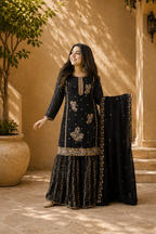 Woman in a black embroidered traditional outfit standing outdoors with a beige wall and potted plant in the background.